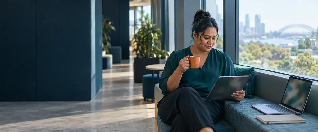 A woman reads a tablet while holding a coffee cup at an office window bench, with a laptop and notebook beside her