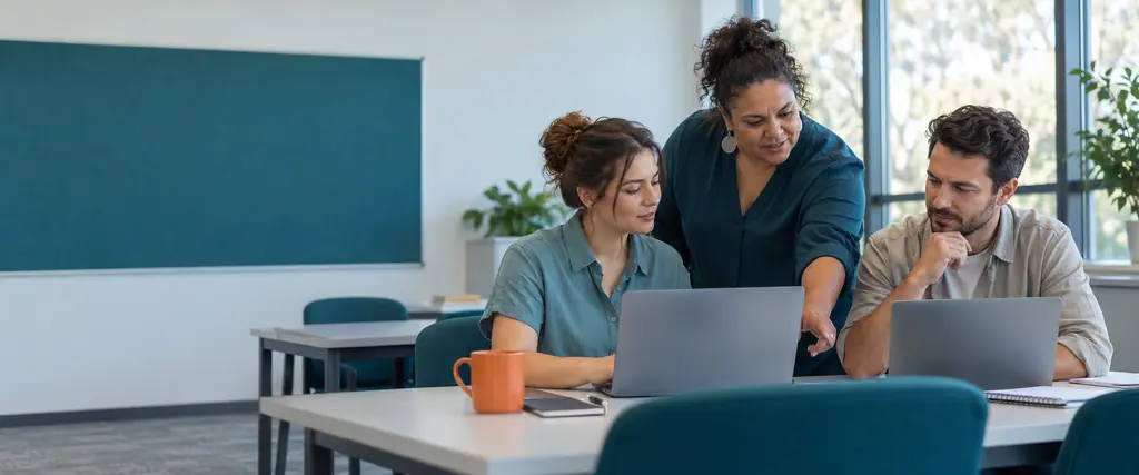 A trainer helps two adult students working on laptops in a bright classroom with a blank teal noticeboard