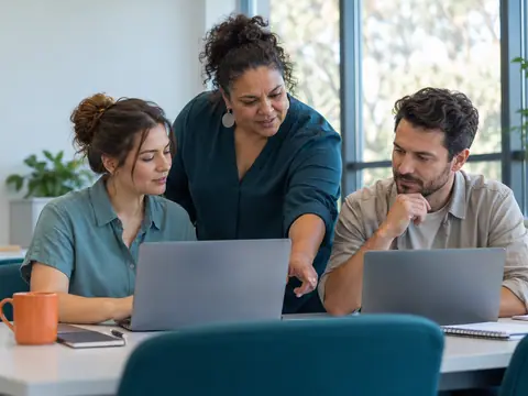 A trainer helps two adult students working on laptops in a bright classroom with a blank teal noticeboard