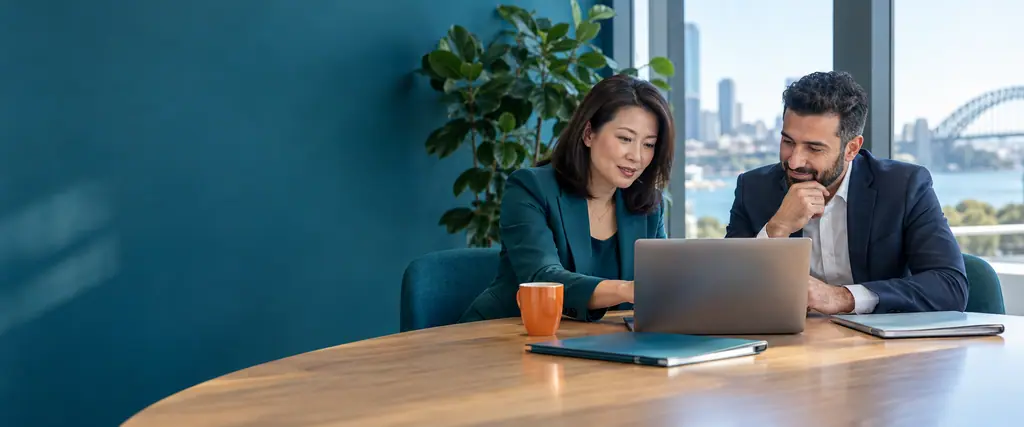 An accountant reviews a laptop with a client at a meeting table in a bright office with a teal wall and plant