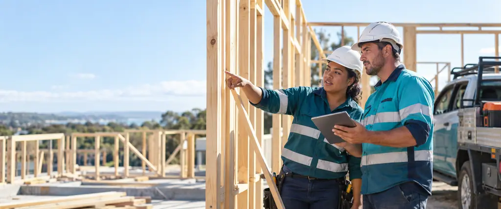 A builder holds a tablet as a woman electrician points at timber framing on a sunny building site, with a ute behind them