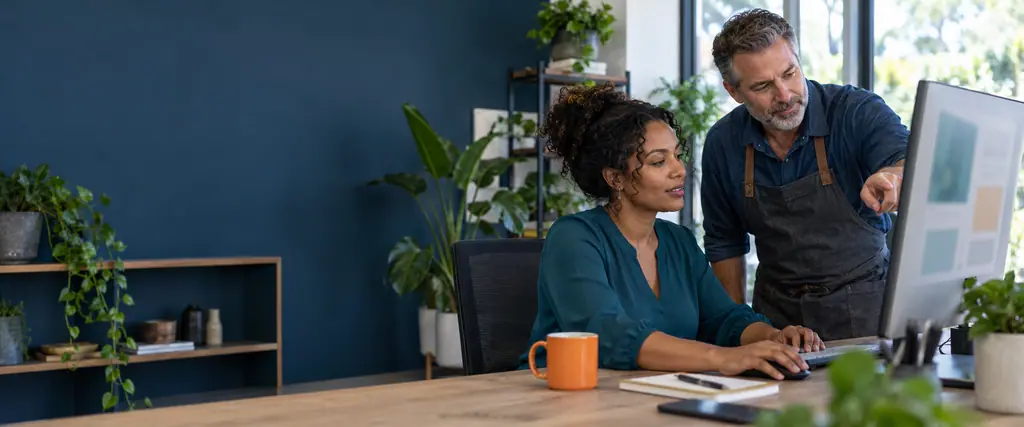 A web designer and an apron-wearing business owner review a blurred page layout on a monitor in a bright, plant-filled office