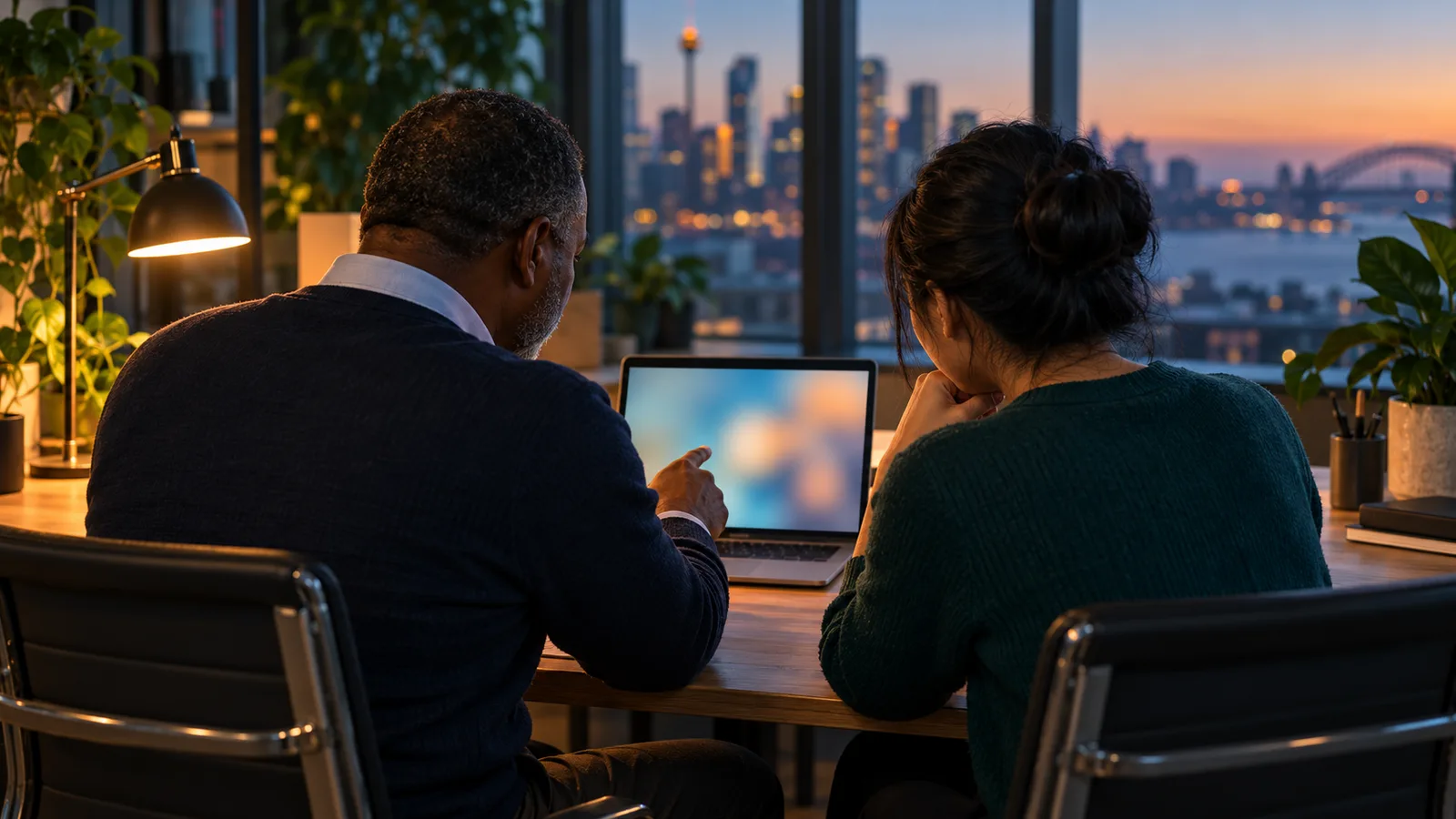 A manager and a young trainee review a laptop together in a teal-toned office lit by a warm desk lamp at dusk.